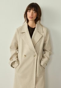 Abrigo de mezcla de lana beige de doble botonadura con amplias solapas, bolsillos frontales y cierre de botón. Textura suave con un ajuste entallado.
