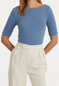 Donna che indossa un top azzurro aderente a maniche corte infilato in pantaloni a vita alta di colore crema con dettagli a bottoni.