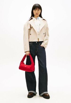 Kvinde iført en beige cropped trenchcoat, hvid bluse, vide mørke jeans, brune sko og holder en rød håndtaske foran en ensfarvet baggrund.