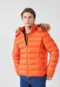 Junger Mann in einer leuchtend oran gefärbten Pufferjacke mit einem mit Pelz besetzten Kapuzen, der vor einem schlichten hellen Hintergrund steht, mit den Händen in den Taschen.