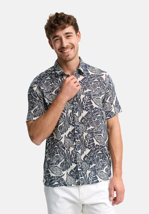 Uśmiechnięty mężczyzna o kręconych włosach poprawiający kołnierz granatowo-białej koszuli w krótkie rękawy z liściastym wzorem, ubrany w białe spodnie, stojący na jednolitym tle.