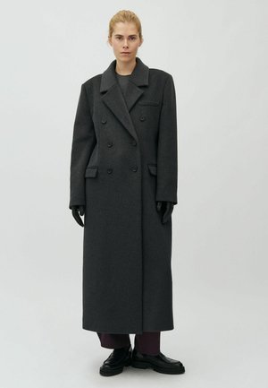 Femme aux cheveux blonds portant un long manteau croisé gris foncé, des gants noirs, un pantalon sombre et des chaussures noires sur un fond clair uni.