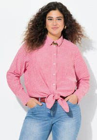 Camicia a righe rosse e bianche con maniche lunghe, annodata in vita, realizzata in un tessuto dalla texture liscia. Abbinata a jeans blu.