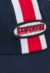Jūras flotes beisbola cepure ar sarkanām un baltām svītrām; satur izšūtu ovālu logotipu ar "tommy" un "Board Sports" kontrastējošās krāsās.