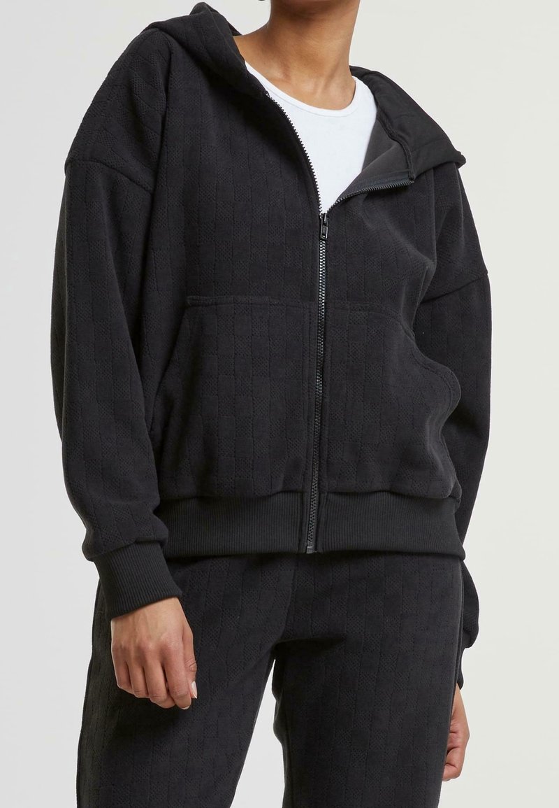 Eine Person trägt einen schwarzen sweatshirtartigen Reißverschluss-Hoodie mit Textur und passende Hosen mit gerippten Bündchen über einem weißen Shirt und posiert vor einem schlichten Hintergrund.