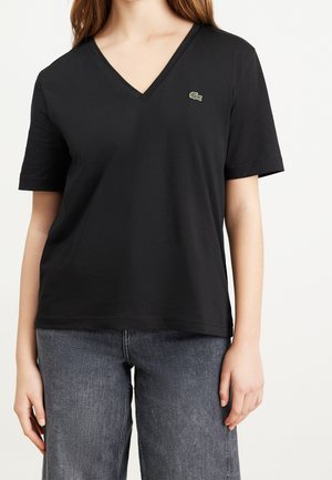 Woman wearing a black V-neck T-shirt with short sleeves and a small green crocodile logo on the left chest, paired with gray denim jeans.
