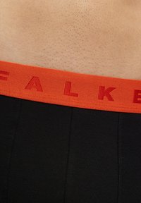 Boxer shorts in tessuto nero con una cintura elastica arancione su cui è intrecciato il marchio "FALKE" in lettere rosse. Texture liscia e design aderente.