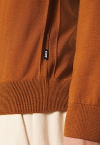 Gros plan sur le bord de manche et l'ourlet d'un pull en maille couleur rouille avec une petite étiquette noire "BOSS", porté sur un pantalon beige clair.