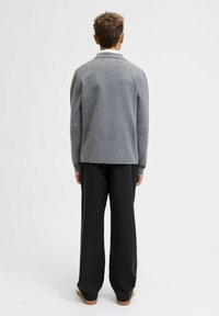 Osoba stoji okrenuta leđima, noseći sivi džemper preko bele košulje, crne pantalone i smeđe cipele na ravnoj beloj pozadini.