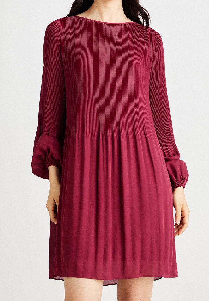 Femme portant une robe bordeaux plissée mi-longue à manches longues avec des poignets froncés et un décolleté bateau sur un fond uni.