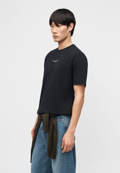 Νεαρός άνδρας που φοράει μαύρο κοντομάνικο μπλουζάκι και μπλε τζιν με σκούρο πράσινο πουλόβερ δεμένο γύρω από τη μέση, στέκεται πλάγια.