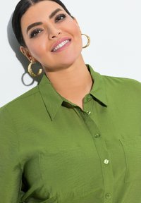Camicia verde con bottoni e colletto, due tasche frontali e un tessuto strutturato. Il modello indossa grandi orecchini a cerchio dorati.