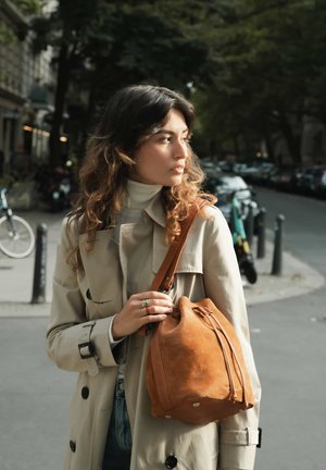 Brown suede bucket bag with drawstring closure and silver hardware, held by a person in a beige trench coat, in a city setting.