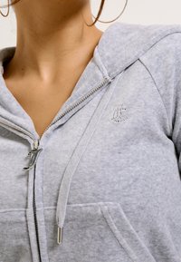 Siva zip-up dukserica od mekog materijala, s velikom kapuljačom, prednjim džepovima i srebrnim patentnim zatvaračem s karakterističnom drškom. Mali detalj s logotipom.