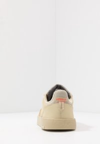 Beige tenisky s texturovaným svrškem a síťovinou, s černobílým puntíkovaným podšíváním a výrazným červeným logem značky na patě.