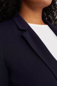 Námořnický blazer s hladkým, texturovaným povrchem. Má úzké klopy a strukturovaný tvar, oblečený přes jednoduchý bílý top.