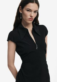 Rochie scurtă neagră cu mânecă scurtă, cu un fermoar în față, guler pliat și detalii plisate pe laterale. Prezintă un accent cu logo metalic aproape de fermoar.