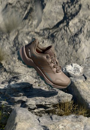 Brun Adidas Terrex vandresko ophængt over stenet terræn med små planter og støvpartikler i luften.