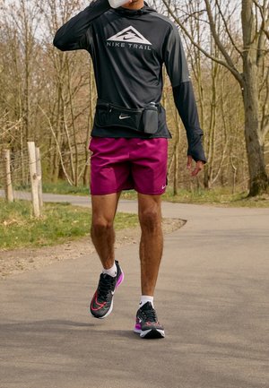 Biegacz w czarnej bluzie Nike Trail, magentowych szortach i czarnych butach do biegania trenuje na utwardzonej ścieżce przez zalesiony obszar.