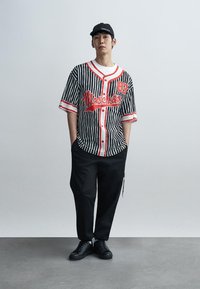 Μπλούζα με κοντά μανίκια σε ασπρόμαυρες ρίγες με κόκκινες λεπτομέρειες και το γράμμα "Moschino", συνδυασμένη με μαύρο παντελόνι και μαύρα αθλητικά παπούτσια.