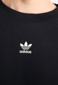 Svart bomullströja med en liten vit Adidas-logotyp med tre blad och horisontella ränder, placerad på bröstområdet.