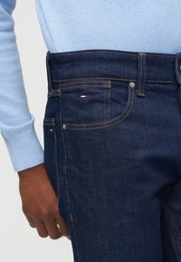 Άνδρας που φοράει σκούρο μπλε τζιν με πορτοκαλί ραφή και ανοιχτό μπλε μακρυμάνικο μπλουζάκι, με το χέρι ακουμπισμένο δίπλα στο ισχίο.