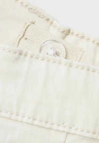Tissu beige avec des coutures visibles, comportant une étiquette au texture semblable à un maillage et un bouton transparent. Attention à la surface lisse et aux détails.
