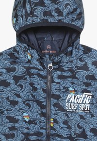 Marineblaue Kapuzenjacke mit hellblauen Wellenmustern, bunten Surfbrett-Icons und weißem Text "Pacific Surf Spot wartet auf die perfekte Welle."