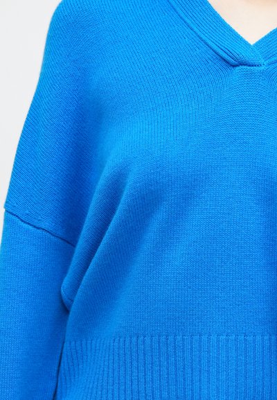 Sweter z długim rękawem w kształcie V w jasnoniebieskim kolorze, z prążkowaną strukturą i luźnym krojem, z wyraźnie podkreślonymi ściegami przy dekolcie i dolnym wykończeniu.