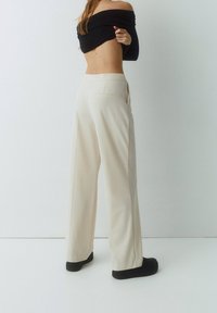 Brede beige broeken met een zachte textuur en achterzakken, gecombineerd met een zwarte off-shoulder top. Zwarte schoenen zijn gedragen.