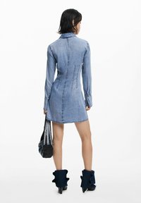 Vestido de mezclilla con mangas largas, cuello y diseño ajustado. Presenta un dobladillo a mitad de muslo y detalles de mezclilla con volantes en las botas hasta el tobillo.