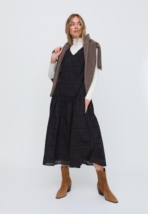 Frau trägt ein schwarzes Midikleid mit floralen Ausschnitten, weißen Rollkragenpullover, braunen Pullover über den Schultern drapiert und beige Wildleder-Stiefeletten.