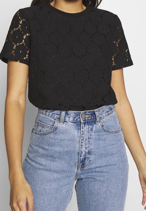 Kobieta ubrana w czarny koronkowy top z krótkim rękawem i wzorami w kształcie kół, do tego jasnoniebieskie, wysokie jeansy z denimu.