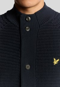 Maglione lavorato a maglia blu navy con design texture, collo alto e tre bottoni neri; presenta un'emblema d'aquila dorata sul petto.