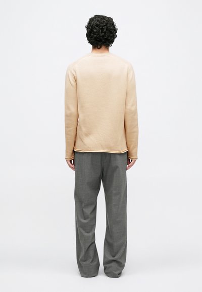 Personne aux cheveux bouclés portant un pull beige et un pantalon gris ample, debout face à un fond blanc uni.