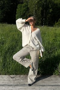 White knitted cardigan with a loose fit and puffed sleeves, paired with beige striped wide-leg pants. Standing on a wooden surface.