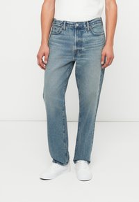 Jeans de mezclilla azul claro con corte recto, cinco bolsillos y detalles de costura. Llevados con zapatillas blancas y una camiseta blanca lisa.
