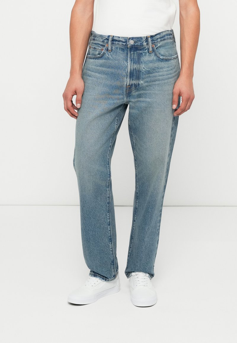 Jeans de mezclilla azul claro con corte recto, cinco bolsillos y detalles de costura. Llevados con zapatillas blancas y una camiseta blanca lisa.