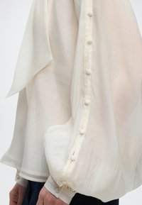 Διαφανές λευκό μανίκι μπλούζας με κουμπιά καλυμμένα με ύφασμα κατά μήκος του εξωτερικού βραχίονα και της μανσέτας, φορεμένο πάνω από σκούρο παντελόνι.