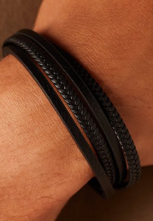 Black leather bracelet with three strands: two smooth and one braided. Textured surface against skin, showing fine details of the material.