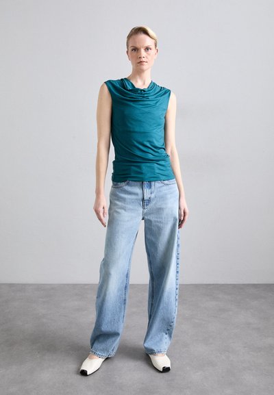 Teal drapert ermløs topp parret med lyseblå vide jeans og hvite sko med svarte tuper, satt mot en nøytral bakgrunn.