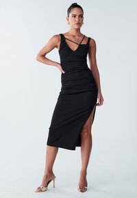 Black fitted dress with ruched detailing, V-neck, and thin cross-strap design. Features a side slit and smooth fabric, complemented by gold heels.