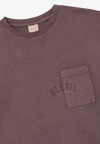 T-shirt z krótkim rękawem w stonowanym fioletowym kolorze, wykonany z bawełny, z kieszonką na piersi i nadrukowanym tekstem "BLKVIS" w ciemnoszarym kolorze nad kieszenią.