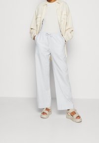 Persoon in een lichtbeige open knoopshirt, wit topje, losse lichtblauwe broek met koord en beige platform sandalen, staande met de handen in de zakken.