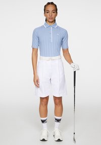 Polo golf azzurro chiaro a maniche corte con un design testurizzato, colletto bianco, abbinato a pantaloni corti bianchi, con una mazza da golf in mano, indossando un guanto e calze sportive.