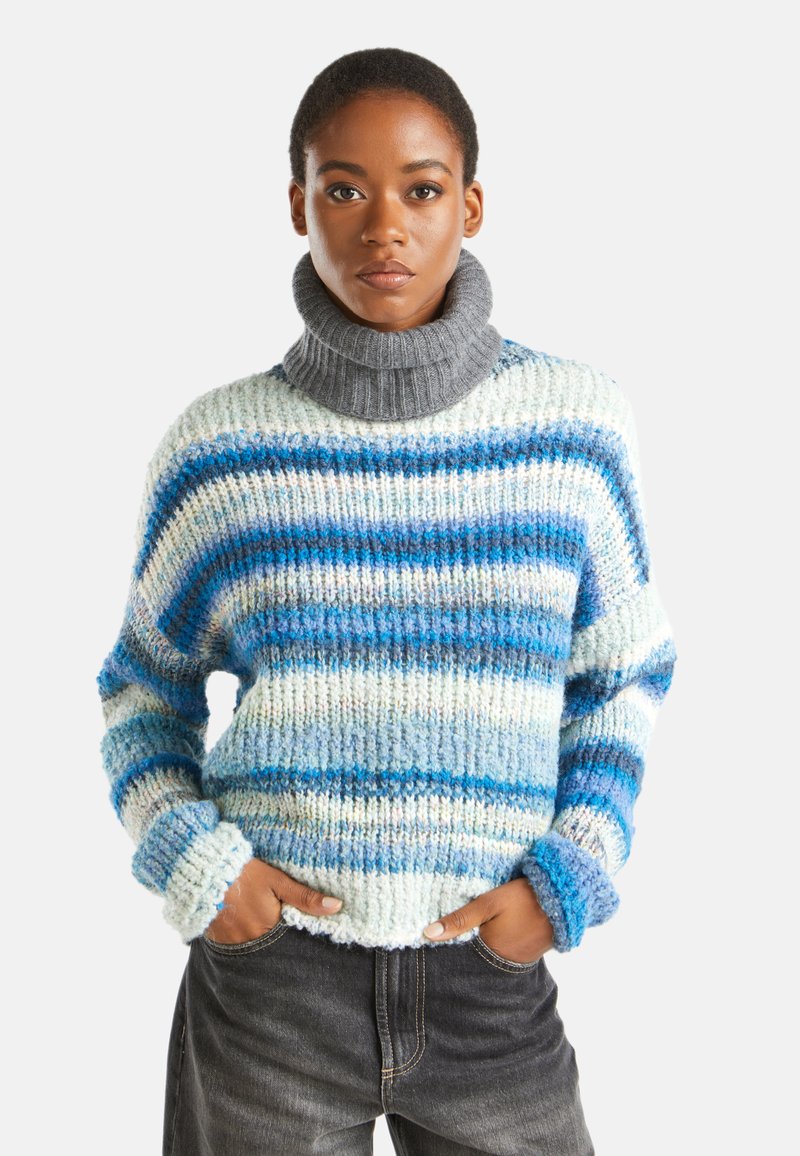 Giovane che indossa un dolcevita grigio e un maglione a righe blu, bianco e grigio, con le mani nelle tasche di jeans neri.