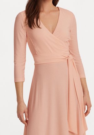 Kobieta ubrana w jasnoróżową sukienkę kopertową z rękawami do łokci i wiązanym paskiem w talii na jednolitym tle.