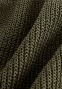 Gros plan sur un tissu en tricot gaufré vert olive montrant un motif quadrillé texturé et en relief ainsi que des plis doux.