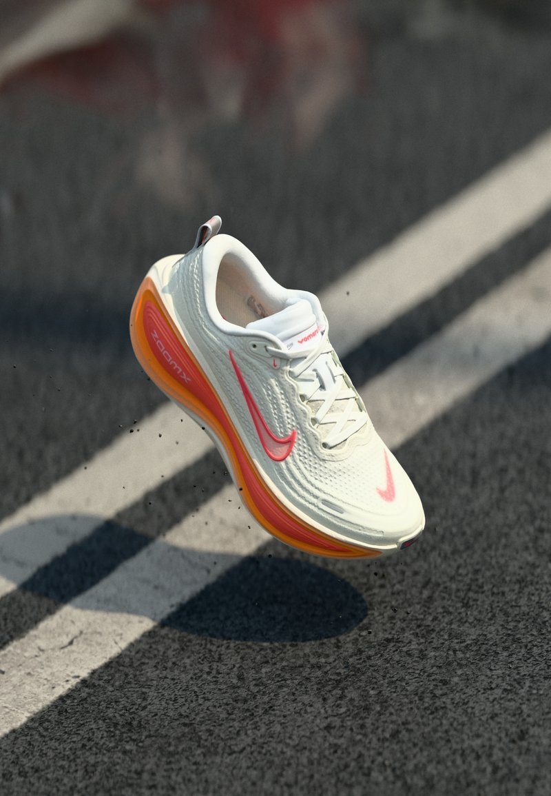 Chaussure de course Nike ZoomX blanche et orange suspendue au-dessus d'une piste avec des marquages blancs.