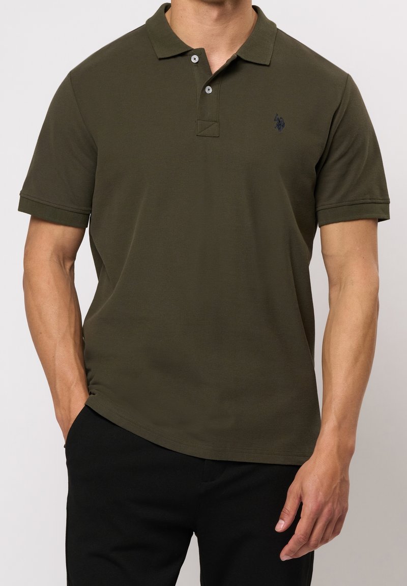 Homme portant un polo à manches courtes vert foncé avec deux boutons et un pantalon noir, debout devant un fond uni.
